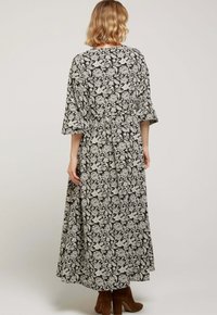 Robe longue noire à motifs floraux blancs ; manches larges, taille froncée et tissu fluide. Portée avec des bottines marron à la cheville.