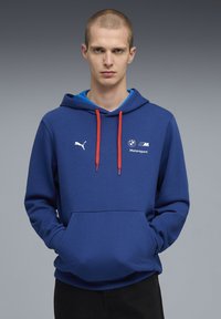 Blauer Kapuzenpullover aus weichem Stoff mit Kängurutasche, roten Kordeln und weißen Puma- sowie BMW Motorsport-Logos auf der Brust.