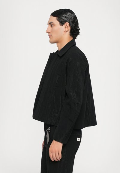 Schwarze croppte Jacke mit strukturiertem Finish, ausgestattet mit einem klassischen Kragen und Seitentaschen. Kombiniert mit schwarzen Hosen und einem Schlüsselanhänger-Detail.