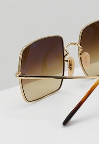 Ochelari de soare cu ramă din metal auriu, cu lentile mari pătrate cu gradient. Perne de nas transparente și un accent pe temple cu model de broască țestoasă.