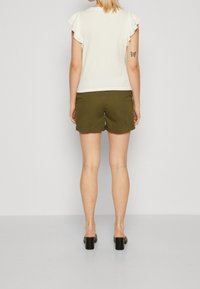 Bílý top s krátkým rukávem a volánkovými rukávy, doplněný olivově zelenými šortkami. Černé boty na blokovém podpatku dotvářejí look.