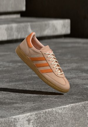Beiger Sneaker mit orangen Streifen, gepolstertem Kragen, strukturierter Gummisohle und weißen Schnürsenkeln. Mit einem "Spezial"-Logo an der Seite.