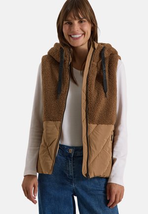 Donna sorridente che indossa un gilet marrone trapuntato e in pile senza maniche con cappuccio sopra una camicia bianca a maniche lunghe e jeans blu.