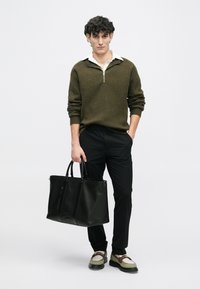 Olivově zelený pletený svetr na zip, černé slim-fit kalhoty, černá kožená kabelka a béžové boty s hnědými akcenty. Neutrální pozadí.