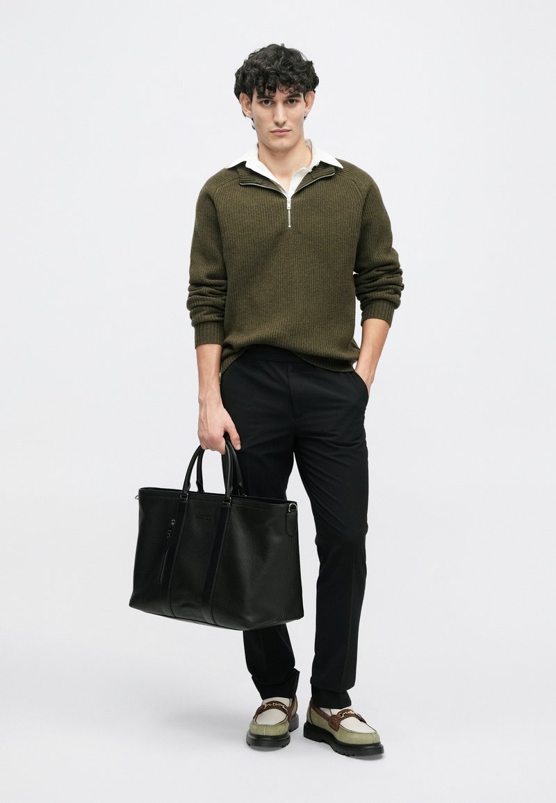 Olivově zelený pletený svetr na zip, černé slim-fit kalhoty, černá kožená kabelka a béžové boty s hnědými akcenty. Neutrální pozadí.