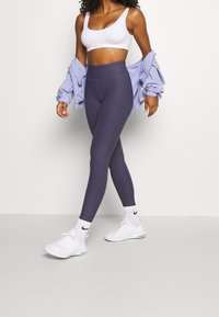 Violetti korkean vyötärön legginssit, joissa on tekstuuripinta ja joustava materiaali, yhdistettynä valkoiseen urheiluliiviin ja vaaleansiniseen takkiin. Valkoiset lenkkarit.