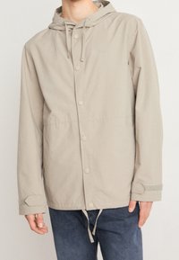 Jasnobeżowa kurtka z kapturem zapinana na napy, z regulowanymi mankietami i sznurkiem w talii noszona z ciemnoniebieskimi dżinsami.