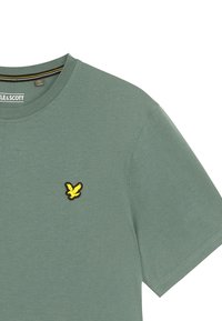T-shirt verde in cotone con scollo rotondo, maniche corte e logo ricamato di un uccello giallo sul petto. Tessuto morbido.