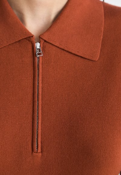Polo en tricot couleur rouille avec une texture lisse, doté d'une fermeture éclair à l'avant et d'un col pointu classique.