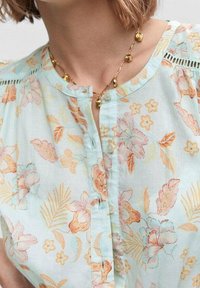 Kwiatowa bluzka z jasnoniebieskiej tkaniny, z pomarańczowymi i różowymi wzorami kwiatów, okrągłym dekoltem, zapięciem na guziki i subtelnymi koronkowymi detalami.