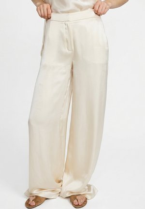 Cremefarbene, weit geschnittene Satin-Hose, getragen mit braunen offenen Sandalen, Hände passen den Bund an, schlichter weißer Hintergrund.