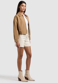 Naine seisab, kandes kõhukest pruuni jakki, valget tippu, kreemikaid lühikesi pükse ja beeže pahkluu saapaid heledal hallil taustal.