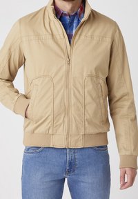 Beige bomber bunda se zipem vpředu, žebrovanými manžetami a lemem, bočními kapsami a hladkým materiálem, nošená přes kostkovanou košili.