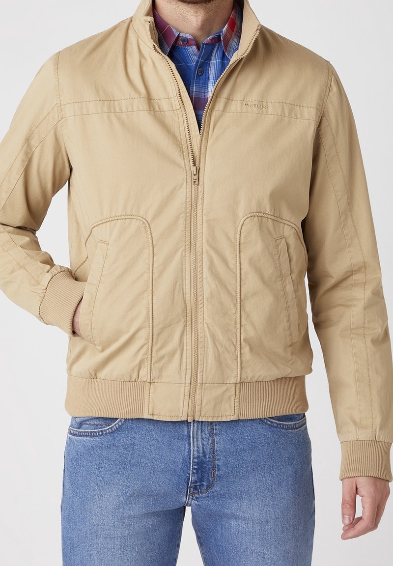 Beige bomber bunda se zipem vpředu, žebrovanými manžetami a lemem, bočními kapsami a hladkým materiálem, nošená přes kostkovanou košili.