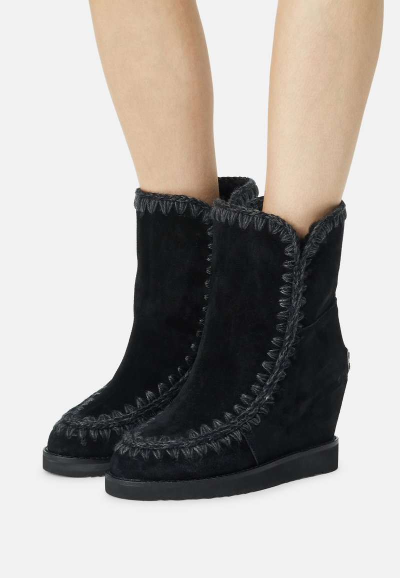 Botas de tacón cuña de ante negro con borde superior en forma de ondas, costuras decorativas y un acabado texturizado. Punta redondeada y suela de goma plana.