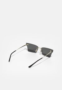 Occhiali da sole neri angolari con lenti scure, sottile montatura in metallo dorato e punte delle aste in plastica nera. Naselli trasparenti per il comfort.