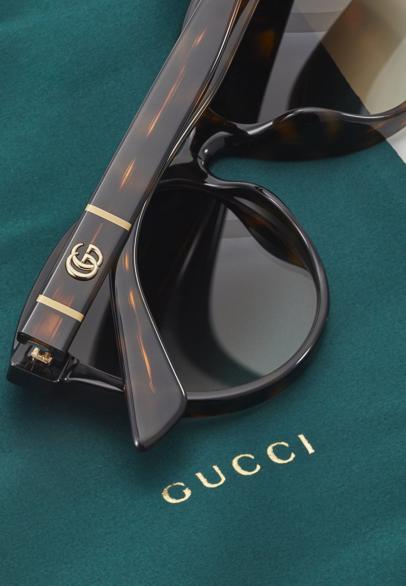 Lunettes de soleil Gucci avec une monture en écaille sombre, des accents en hardware doré et des verres foncés reposant sur un fond en tissu vert.