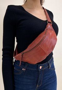 Borsa a tracolla in pelle marrone con una forma curvata, dotata di un vano principale con zip e una tracolla regolabile, indossata sopra un top nero.