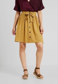 Hame a khukum haírti maiñ maustard rang ki, jis mein elastic waistband, bandhan ka tafaail, aur saamne ke batan hain, jise bhoore strappy espadrille sandals ke saath pehna gaya hai.