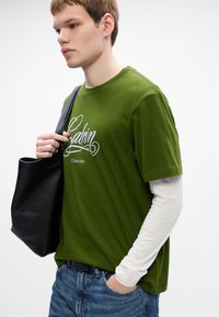 Young man wearing a green Calvin Klein t-shirt over long white sleeves, blue jeans, holding a black shoulder bag, standing against white background.