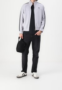 Lichtgrijze vest met ritssluiting en zwarte strepen, een zwart T-shirt, donkere spijkerbroek, witte sneakers met zwarte accenten en een kleine zwarte schoudertas.