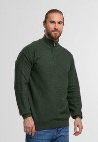 Mann mit Bart und zurückgebundenem Haar, der einen dunkelgrünen Half-Zip-Pullover und blaue Jeans trägt, steht vor einem schlichten, hellen Hintergrund.