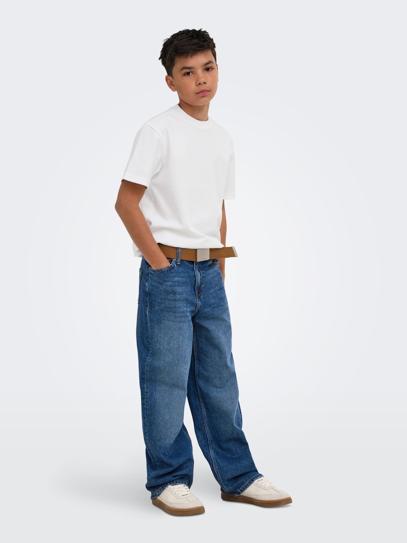 Garçon debout les mains dans les poches, portant un t-shirt blanc, un jean large bleu avec une ceinture marron et des baskets blanches sur fond uni.