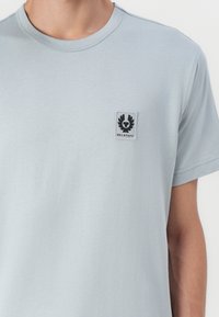 Camiseta de manga curta azul clara, feita de um tecido macio, com um pequeno emblema preto no lado esquerdo do peito. Textura suave e corte descontraído.