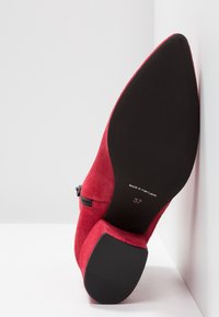 Bottine en daim rouge avec fermeture éclair, bout arrondi et semelle en caoutchouc noire. Taille 37 indiquée sous la chaussure. Contraste de texture net.