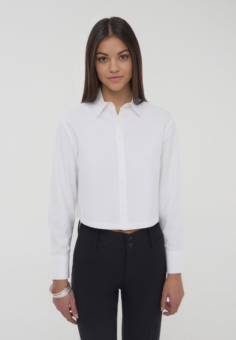Camicia bianca a maniche lunghe con bottoni, dotata di colletto classico e tessuto morbido, abbinata a pantaloni neri aderenti.