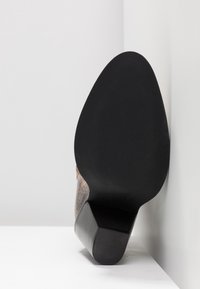 Zapato de tacón alto marrón con un diseño estampado, suela negra y lisa, y tacón en bloque, apoyado en un ángulo contra una pared blanca.