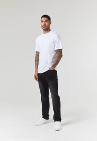 Camiseta blanca de manga corta combinada con jeans negros y zapatillas blancas. El modelo tiene tatuajes y está de pie contra un fondo neutral.