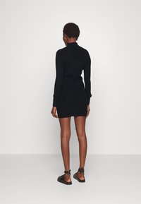 Svart, åtsittande stickad klänning med långa ärmar och hög krage. Har guldfärgad knappdetaljer vid ärmsluten. Kombineras med svarta slip-on skor.