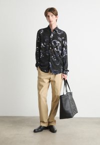 Schwarze Hemd mit Knöpfen und glänzendem Muster, beiges Hosen, schwarze Ledertasche und schwarze Schuhe vor einem neutralen Hintergrund.