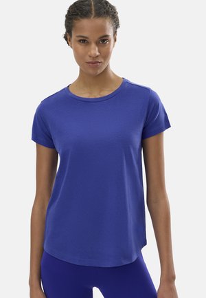 Mujer con el cabello trenzado que lleva una camiseta ajustada de manga corta azul real y leggings a juego, mirando hacia adelante con expresión neutral.