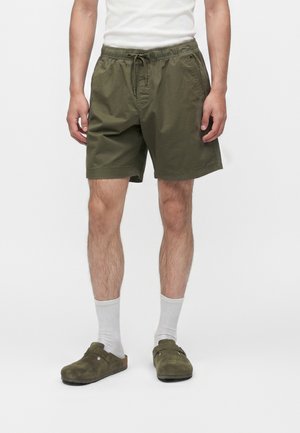 Mann trägt olivgrüne Shorts mit Kordelzug, weiße Socken und olivgrüne Slipper vor schlichtem Hintergrund.