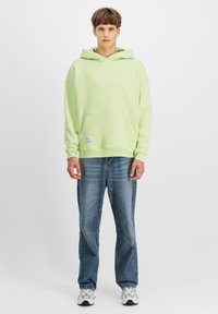 Giovane uomo in piedi che indossa una felpa verde chiaro, jeans blu e sneakers bianche su uno sfondo chiaro e semplice.