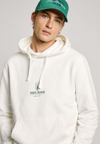 Sudadera blanca con cordones y bolsillo delantero, presenta el logo en verde de "Pepe Jeans". Combinada con una gorra verde que muestra texto bordado.