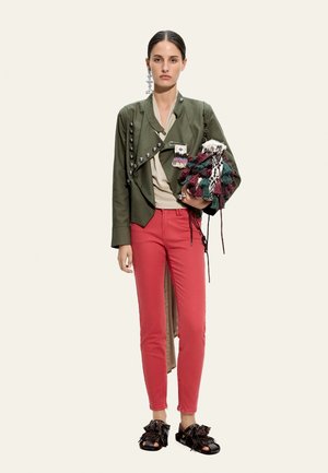 Kobieta w oliwkowej kurtce z metalowymi zdobieniami, beżowym topie, czerwonych spodniach, czarnych sandałach, trzyma wielokolorową torebkę z frędzlami, nosi długie kolczyki.