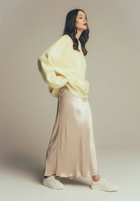 Jasnożółty, oversize'owy sweter, beżowa satynowa spódnica midi i białe trampki. Gładkie tekstury i luźna sylwetka tworzą nowoczesny wygląd.