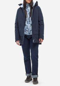 Frau trägt eine dunkelblaue Kapuzenjacke über einem hellblauen gemusterten Reißverschluss-Fleece, dunklen Jeans mit Umschlägen und grauen Wanderschuhen.