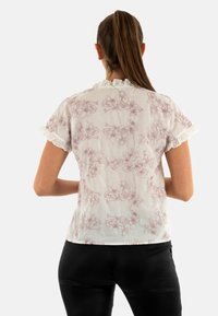 Blusa bianca a maniche corte con un motivo floreale in rosa chiaro, caratterizzata da bordi arricciati su maniche e scollatura. Tessuto morbido e leggero.