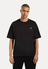 T-shirt in cotone nero con collo rotondo e maniche corte, caratterizzato da un piccolo etichetta rettangolare bianca sul lato sinistro del petto.