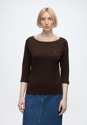 Femme aux cheveux blonds courts portant un haut marron foncé à manches longues et une jupe en jean bleue, debout devant un fond blanc uni.