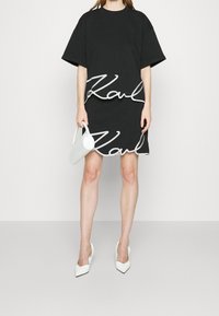 Černá bavlněná oversized tričko a sukně s výrazným bílým nápisem "Karl". Modelka drží bílou tašku a má na sobě bílé lodičky.