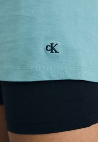 Light blue cotton t-shirt with short sleeves, featuring a navy embroidered "cK" logo near the hem, paired with black fitted shorts.