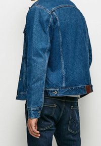 Giacca in denim blu scuro con vestibilità comoda, cuciture visibili, dettagli con bottoni e una patch in pelle sul retro. Design classico con tasche laterali.