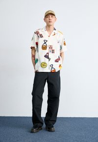 Camisa de manga corta color crema con gráficos retro coloridos. Pantalones negros, zapatos negros con suela en contraste. Una gorra simple completa el conjunto.