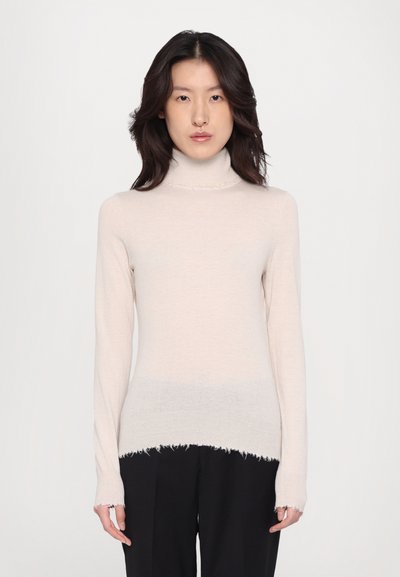 Pull à col roulé beige clair avec des manches longues, présentant des détails de bord frangé et une texture douce et lisse. Associé à un pantalon noir.