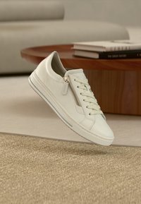 White leather sneaker featuring lace-up front and side zipper, textured sole, and subtle gray stripe. Floating above a textured surface.